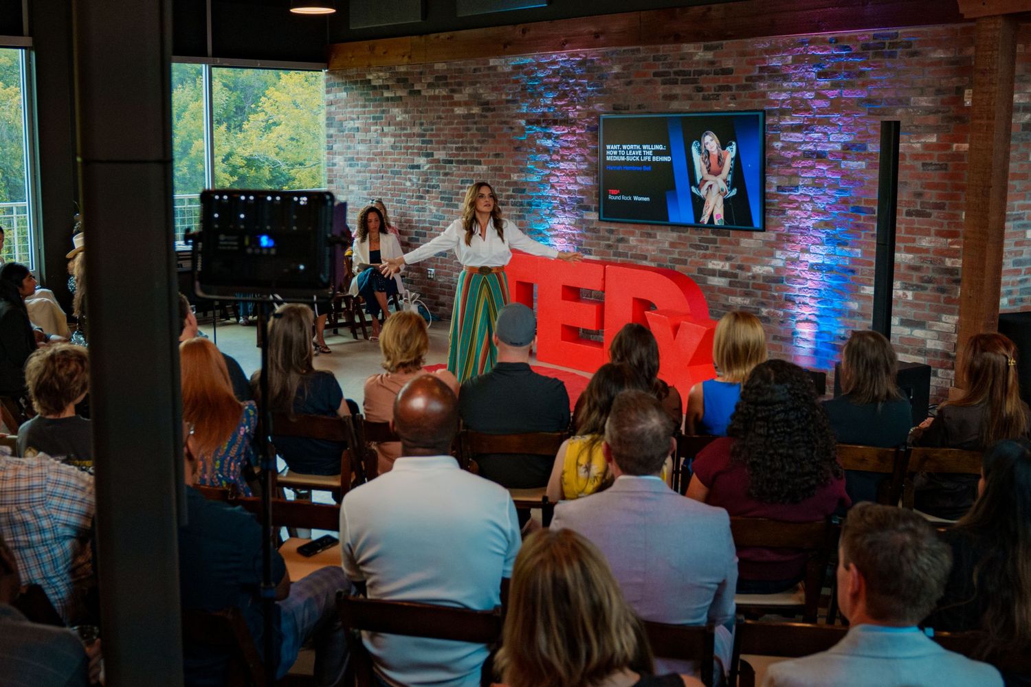 Hannah Hembree Bell  speaks on stage at a TEDx event before a seated audience in a brick-walled venue with red TEDx logo. Round Rock Texas.