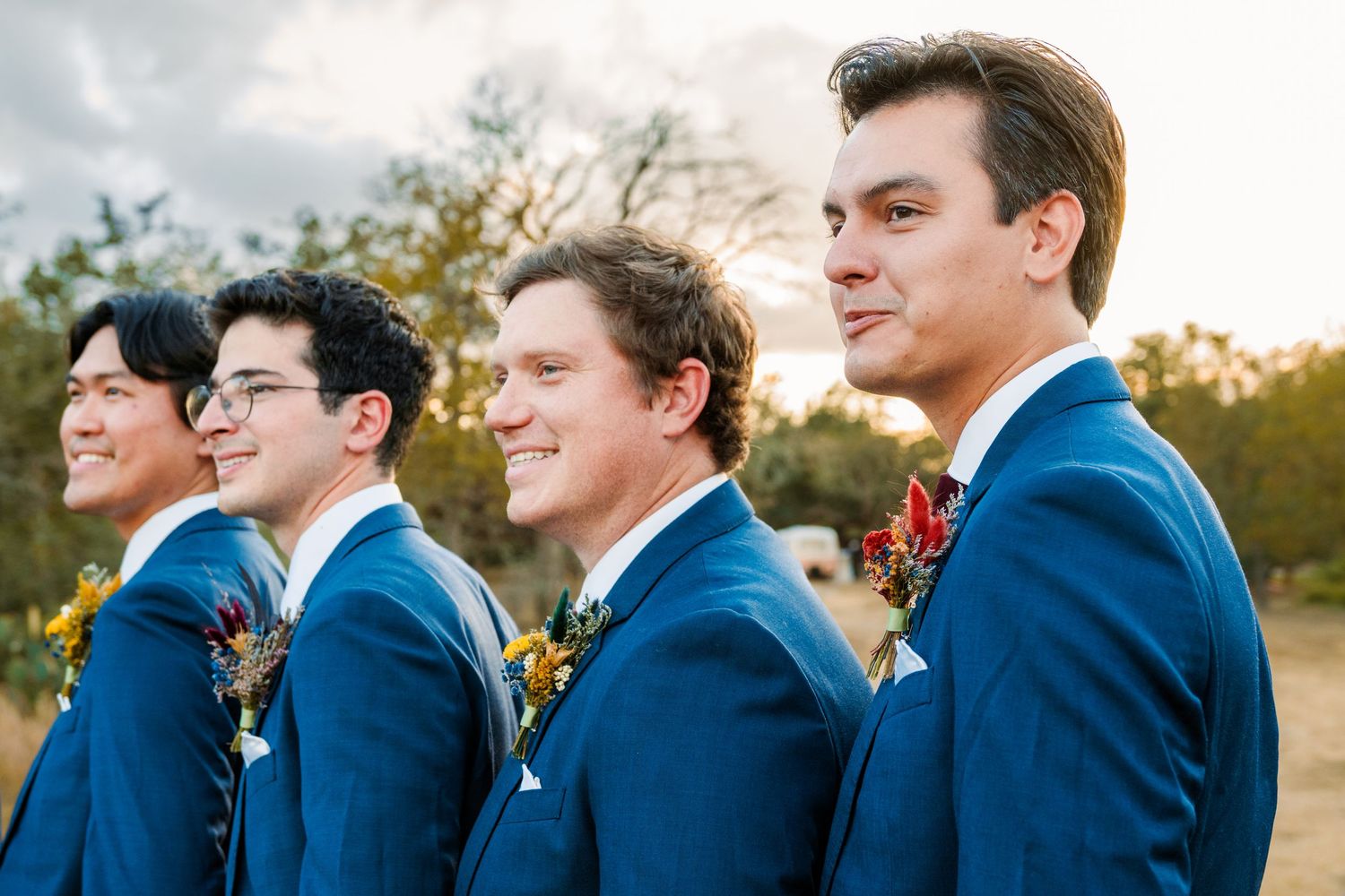 Four groomsmen in navy blue suits with floral boutonnieres pose outdoors at a wedding ceremony.