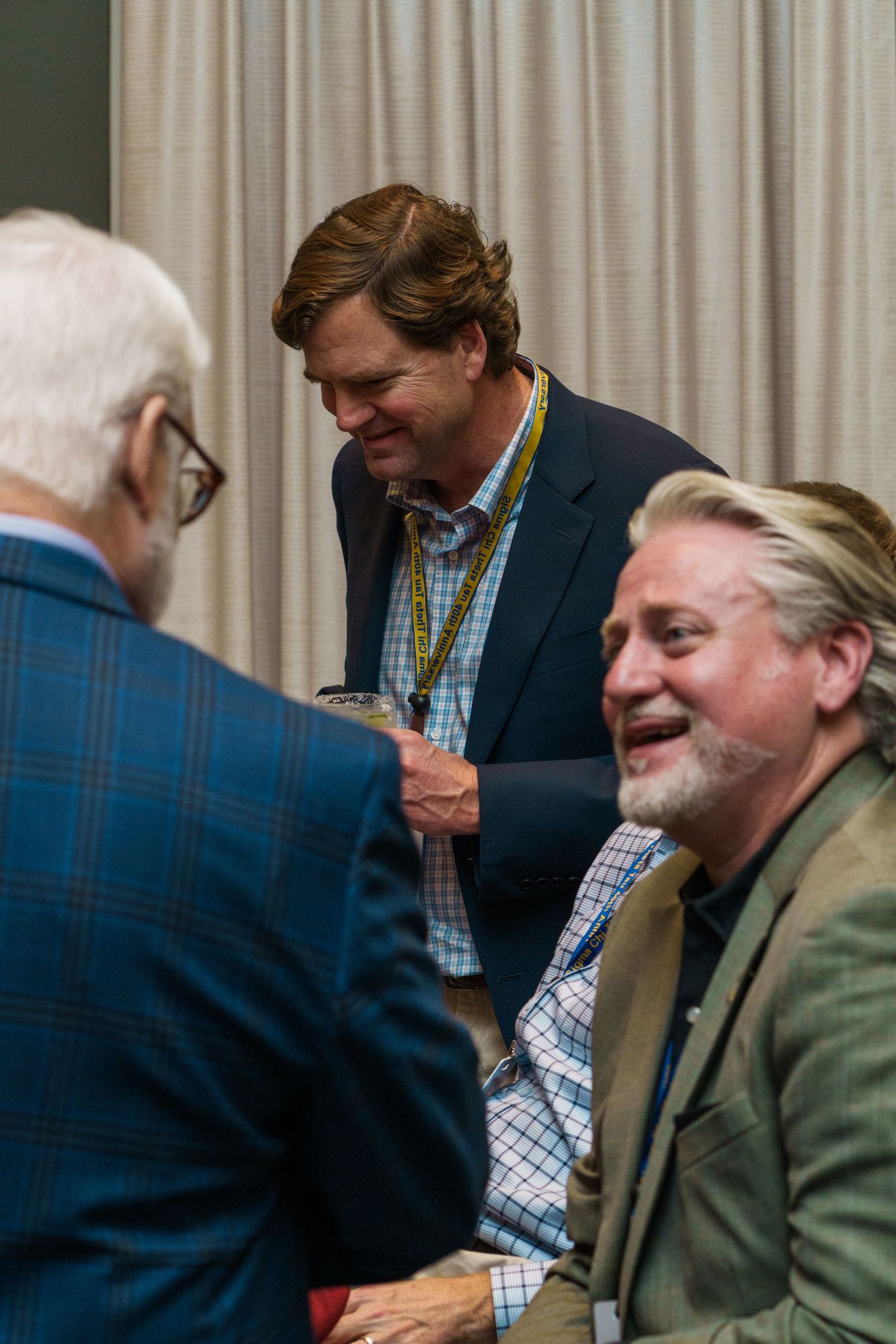 Three men in business casual attire laugh and converse at a professional networking event or conference.