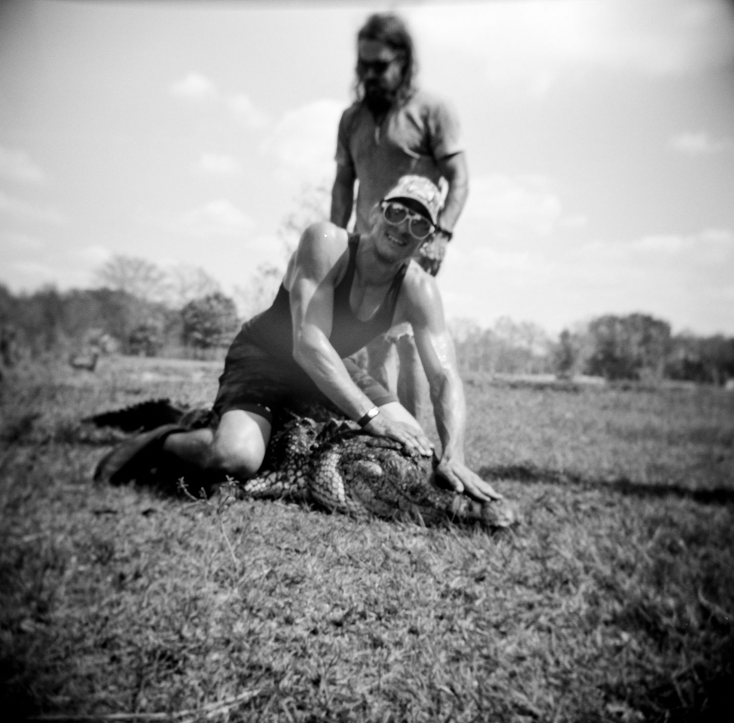 Man in sunglasses pins an alligator to the ground while another person stands behind, black and white.
