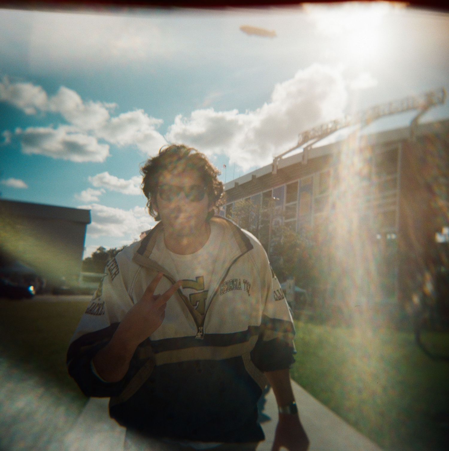 Young man in a racing jacket poses outdoors with lens flare and blue sky behind him.