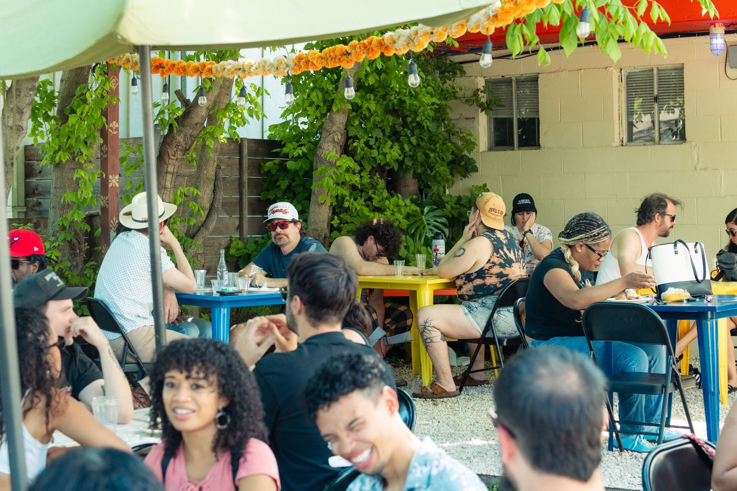 Large group of diverse people dining and socializing at colorful outdoor tables under trees.