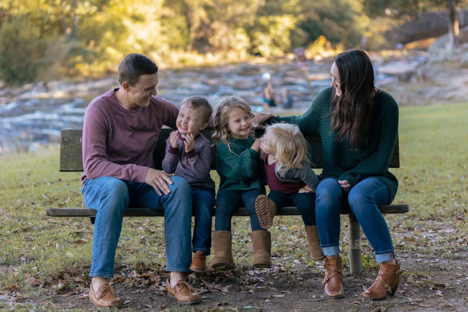 Happy family of five sitting on a park bench together, laughing in autumn sunlight.