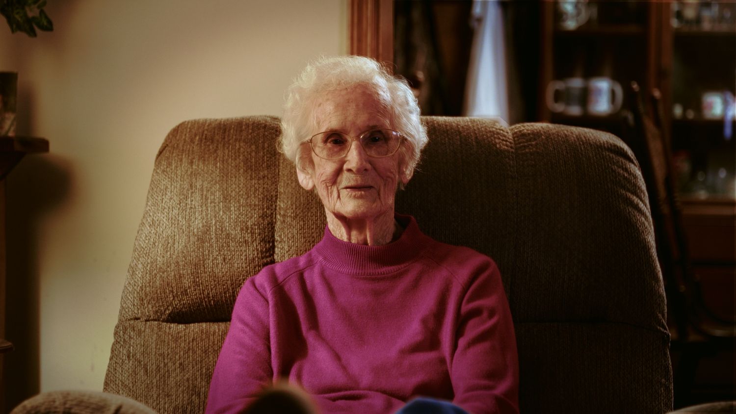 Elderly woman with white hair wearing a pink sweater sitting in a brown recliner chair in a cozy living room.