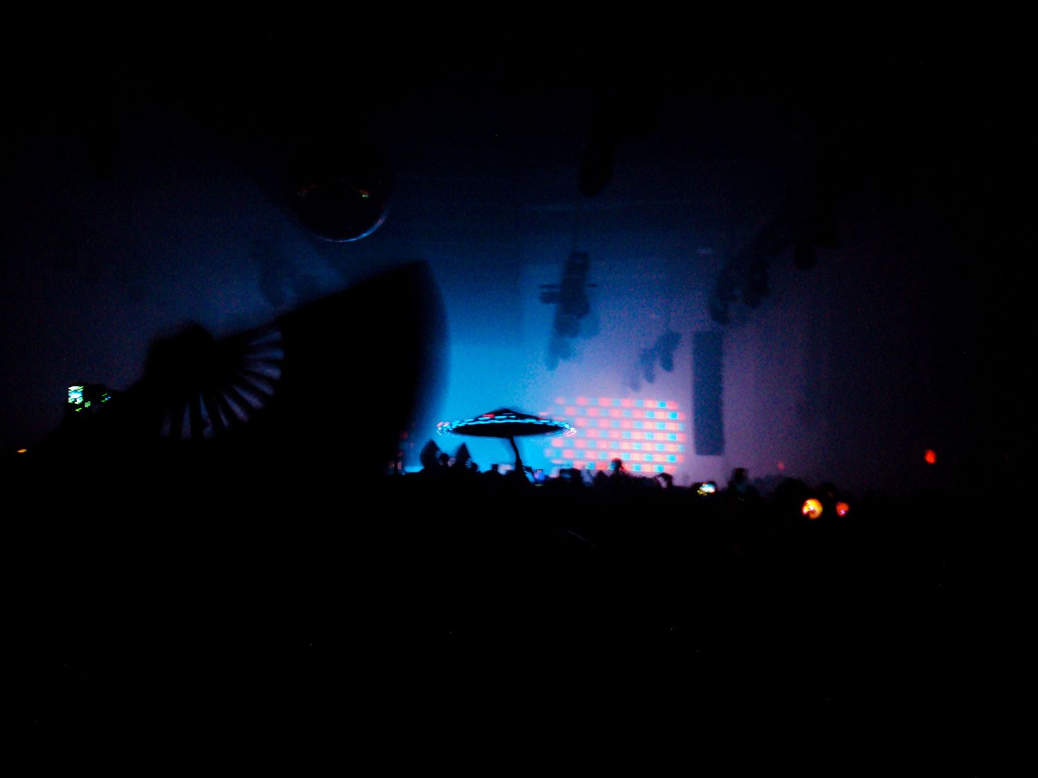 Dark nightclub scene with blue-lit brick wall, silhouetted crowd, DJ umbrella stand, and large shark artwork.