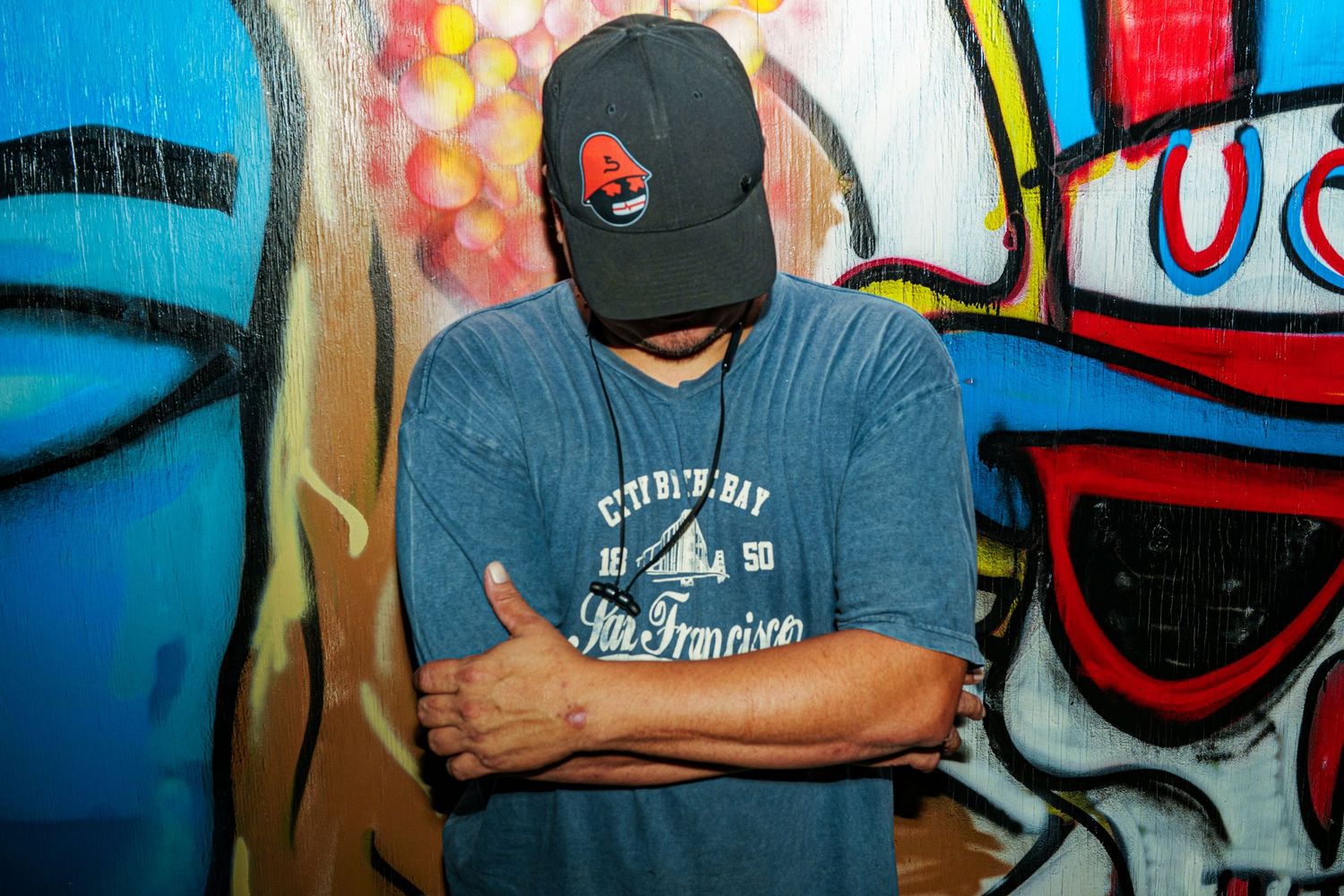 Man in blue San Francisco t-shirt and black cap poses with arms crossed against colorful graffiti wall.