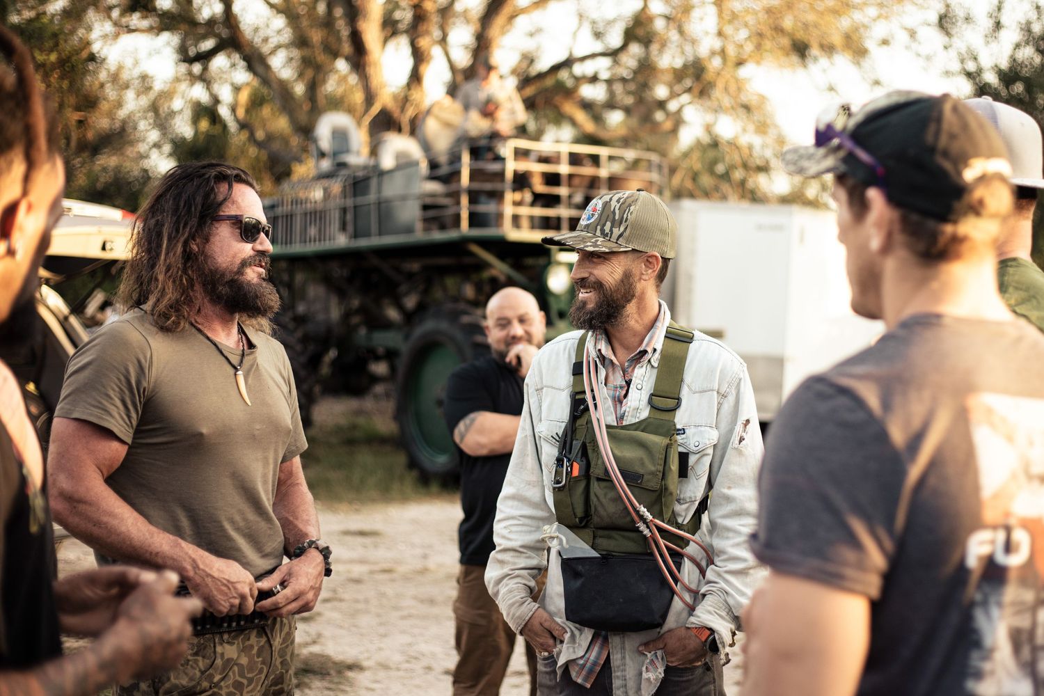 Men in tactical gear conversing on an outdoor film set with a large military vehicle in the background.