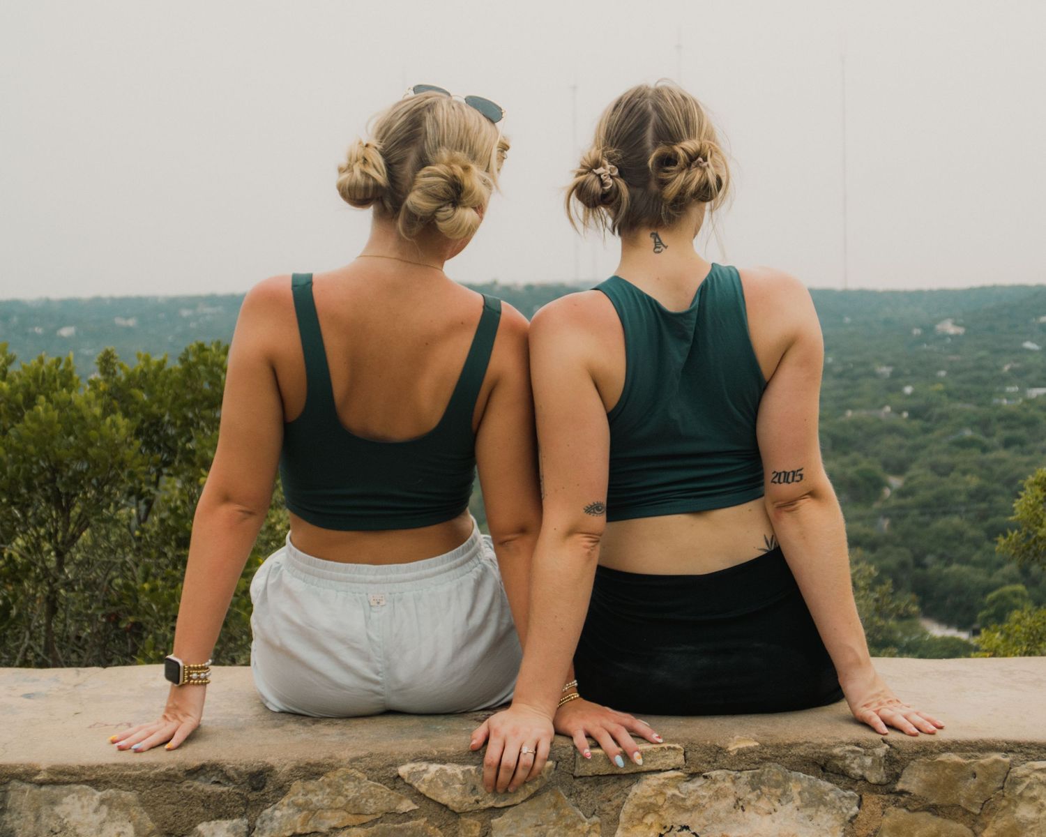 Two women with updo hairstyles wearing teal sports bras sit on a stone wall overlooking a lush green landscape.