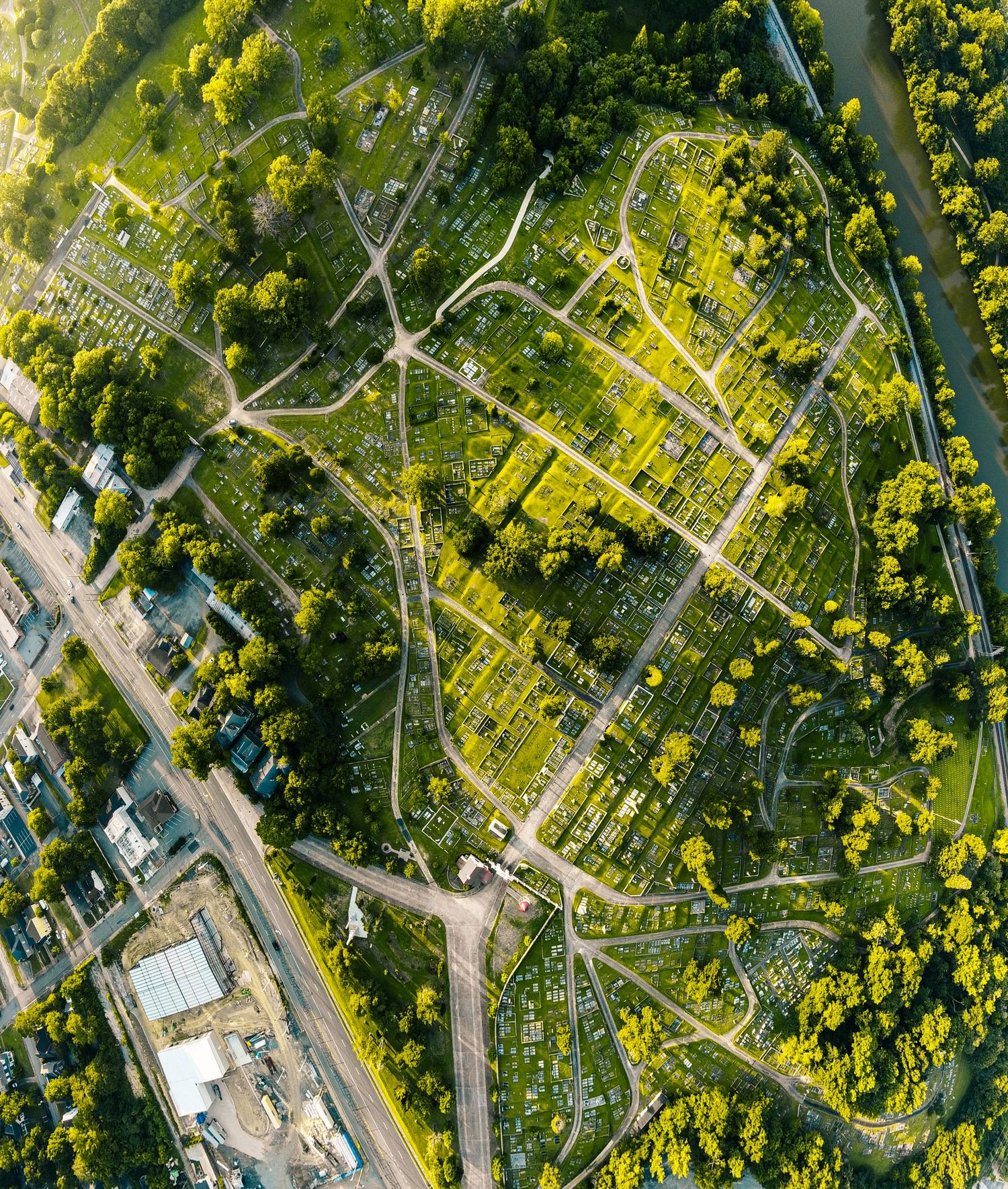 Aerial view of a lush green cemetery with winding paths surrounded by dense trees and urban infrastructure.