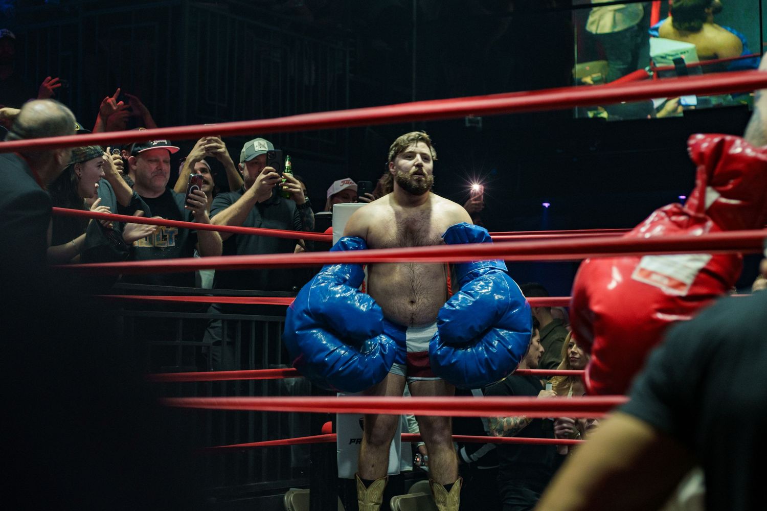 Boxer wearing oversized blue gloves stands between ring ropes while fans photograph the match.