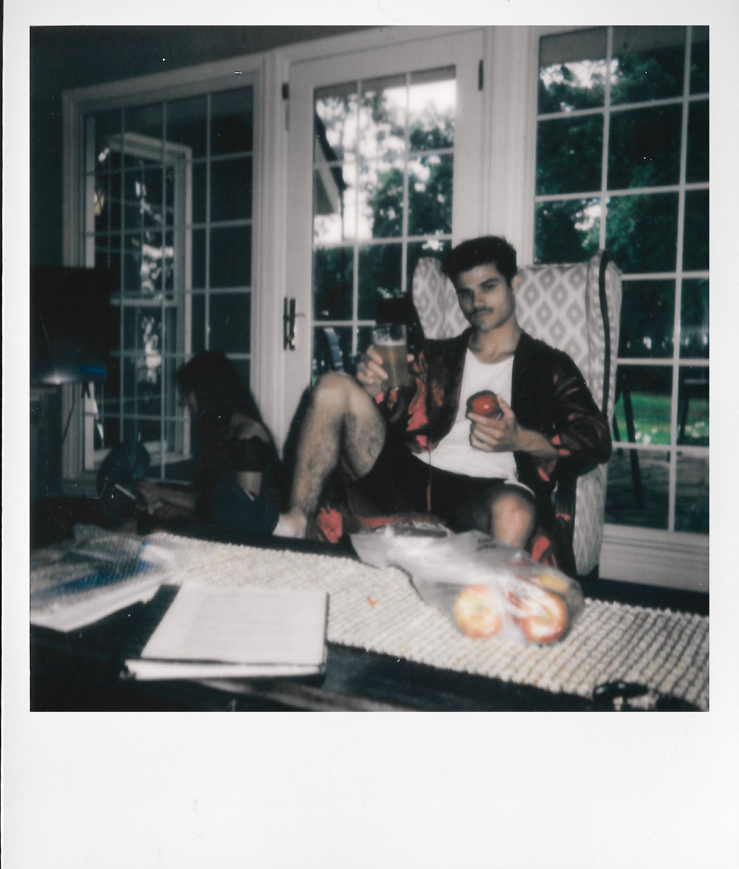Young man relaxing in a chair holding an apple, surrounded by papers and fruit on a table near French doors.