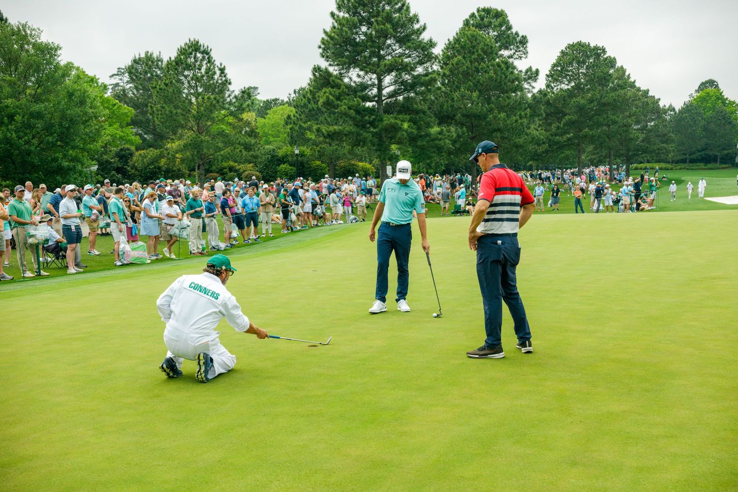 Golfers on a lush green putting green at a Masters Tournament, with a caddie crouching to read the putt before a large crowd.