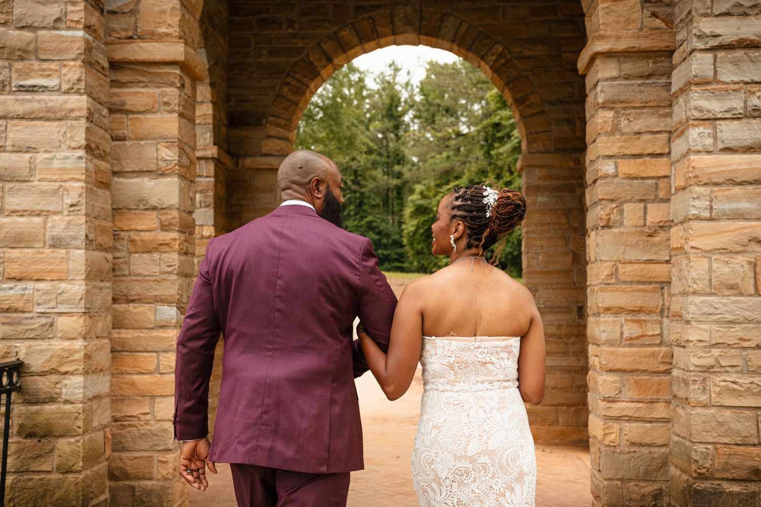Newlyweds walking hand in hand through a stone archway, groom in burgundy suit and bride in lace gown.
