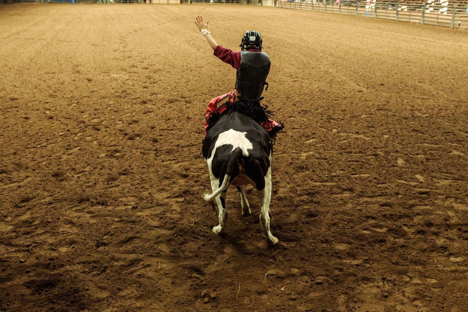 A rodeo rider waves one hand while riding a black and white bull in a dirt arena.