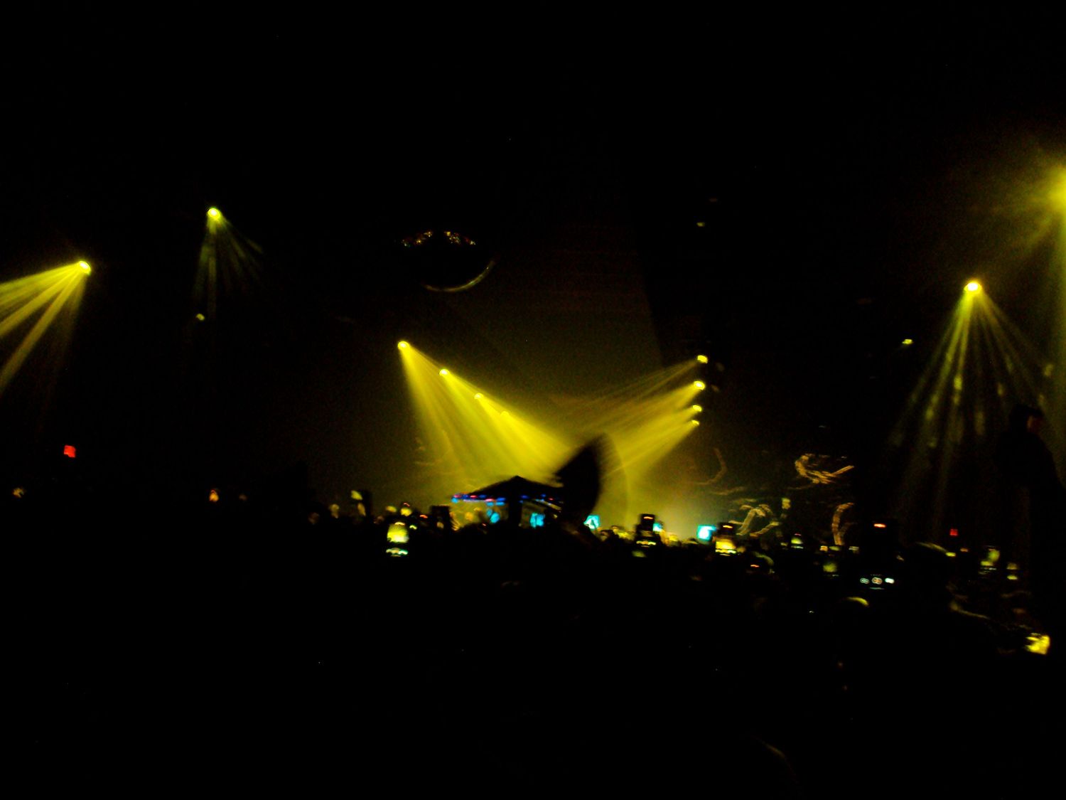 Concert stage with dramatic yellow spotlights illuminating performers and crowd in a dark venue.