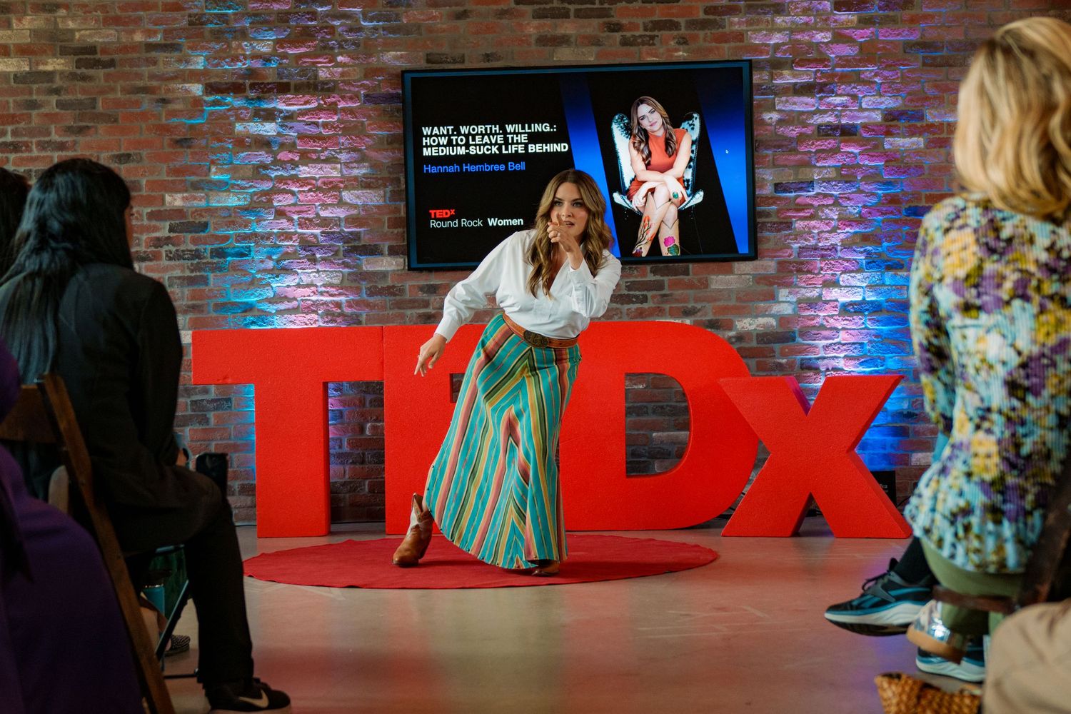 Hannah Hembree Bell presenting on a TED Talk stage with red TEDx letters, brick wall backdrop, and audience members watching, Round Rock TX