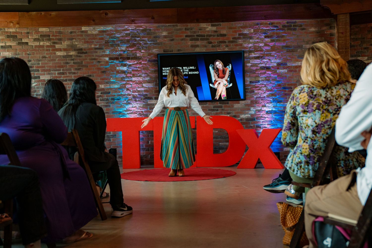 Hannah Hembree Bell presents on a TEDx stage before an audience, wearing a green skirt, with the red TEDx logo visible, Round Rock Texas.