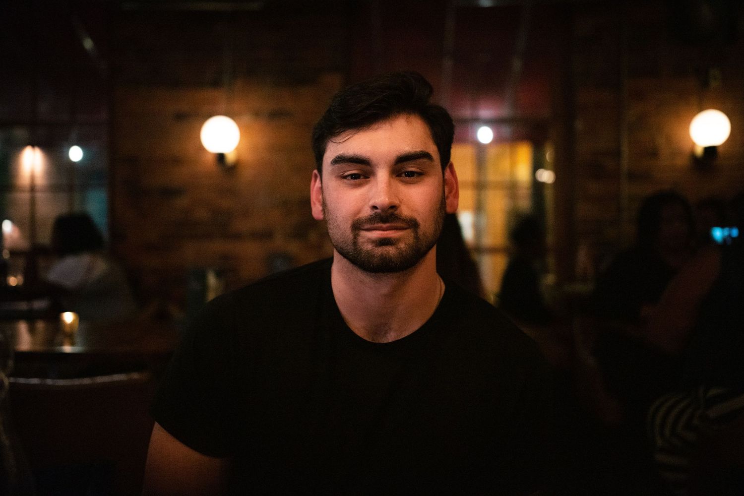 A bearded man with long dark hair wearing a black t-shirt sits in a dimly lit bar with brick walls.