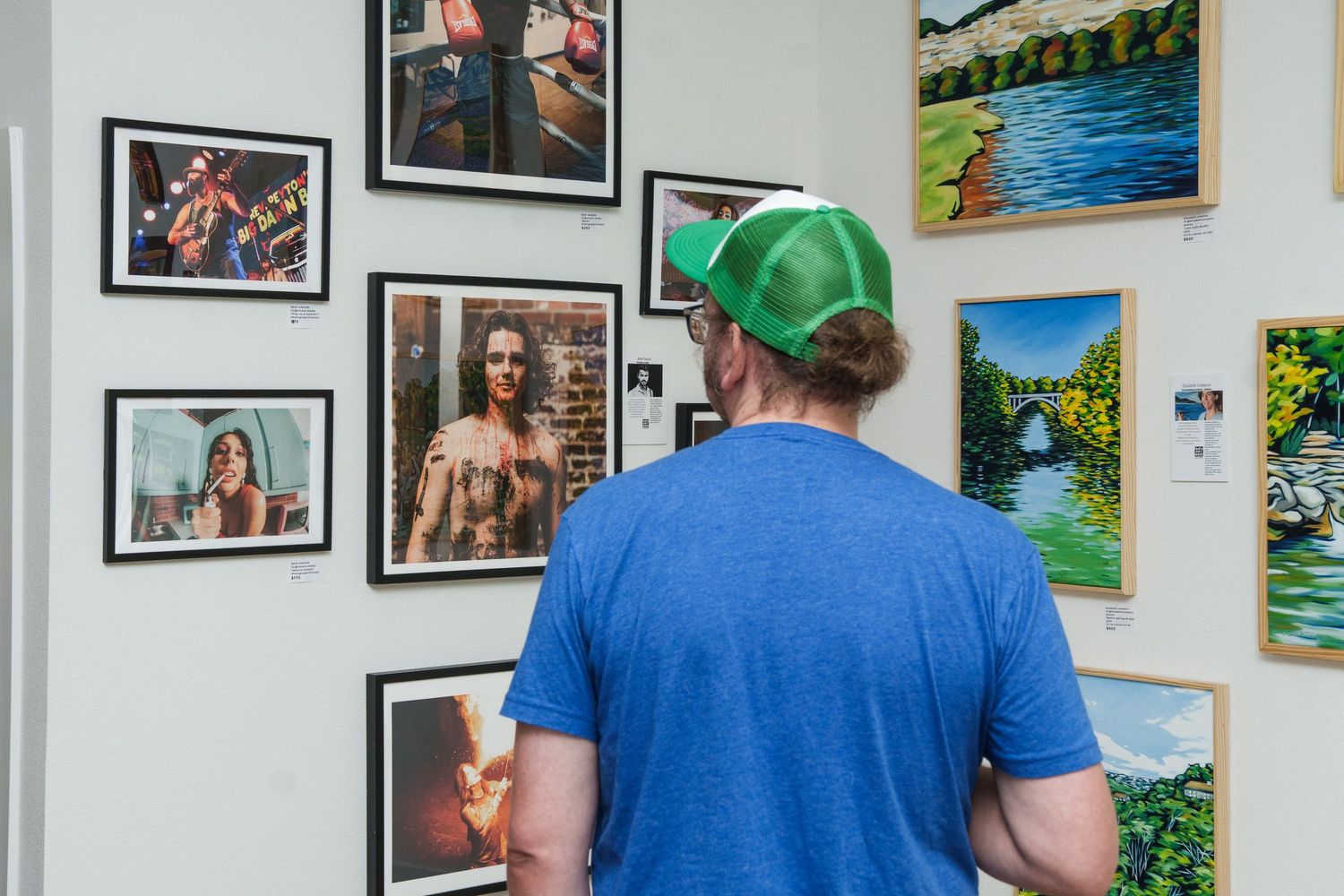 Man in green cap and blue t-shirt viewing framed photos and paintings displayed on a white gallery wall.