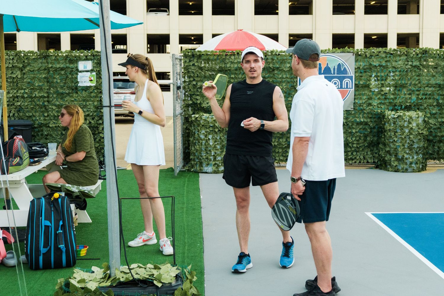 Three people socializing on an outdoor pickleball court, holding paddles, with a green hedge backdrop.