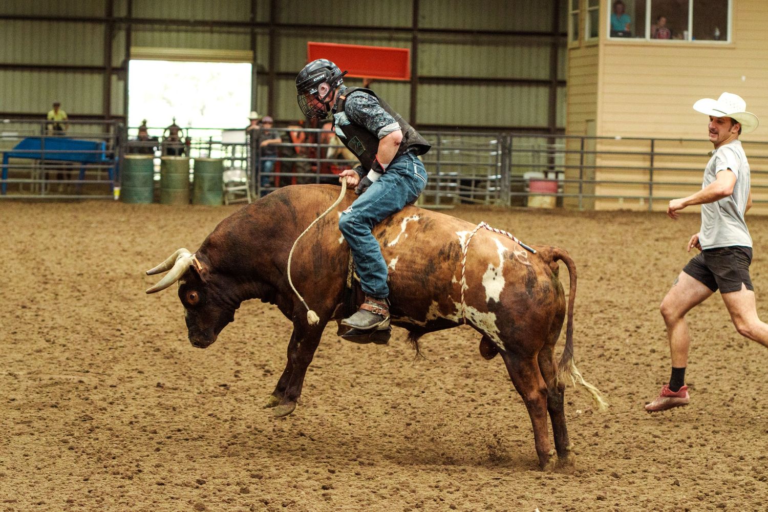 A rodeo rider in protective gear attempts to stay on a bucking brown bull in an indoor arena.