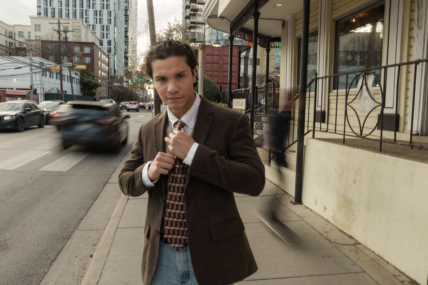 Candid portrait of a young man in a brown suit adjusting his tie while walking along an urban sidewalk, Austin street style.