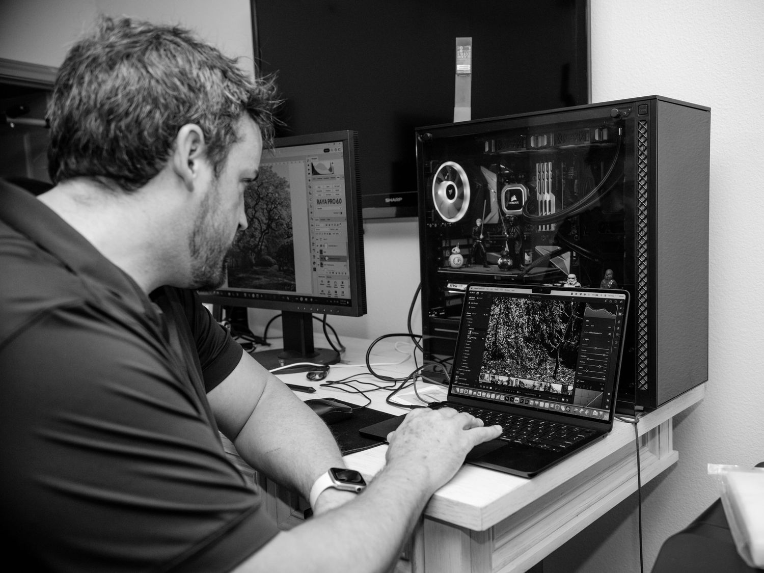 Black and white photo of man editing images on a laptop beside a desktop computer setup.