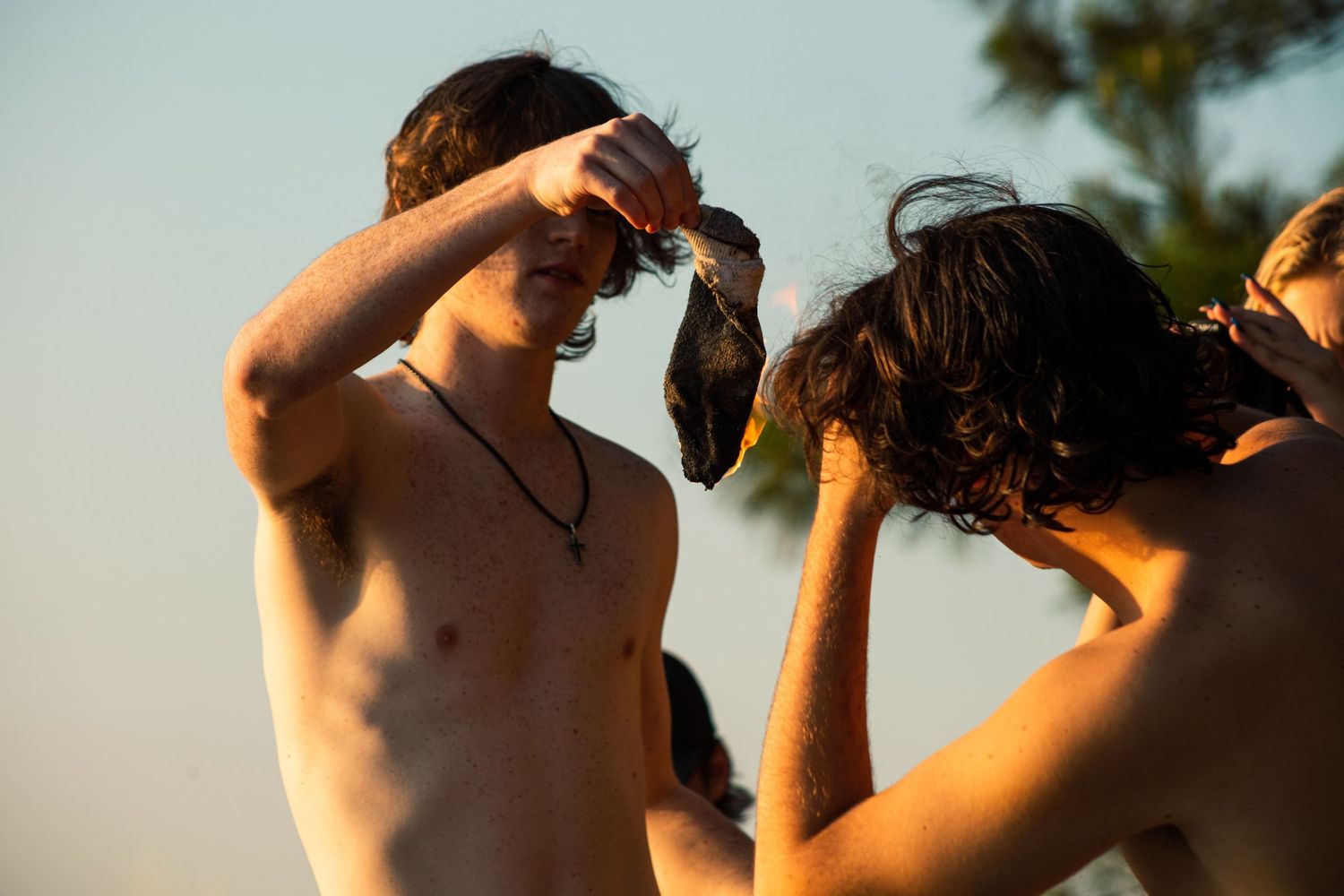 Two shirtless young men outdoors in warm golden light, one holding an object near his head.