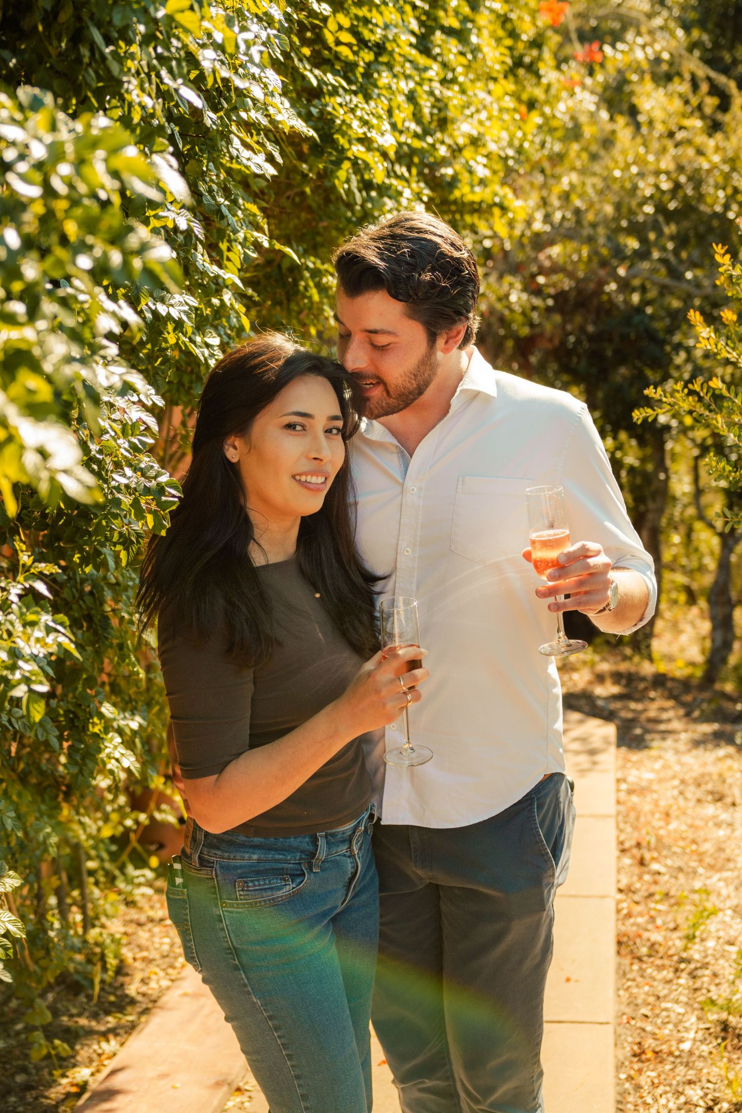 Couple holding wine glasses and embracing in a sunny vineyard, smiling together among lush green vines.