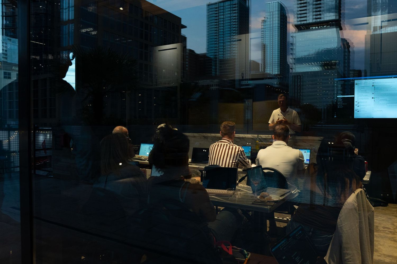 Professionals attend a dimly lit conference room presentation with laptops, city skyline reflecting in glass windows.