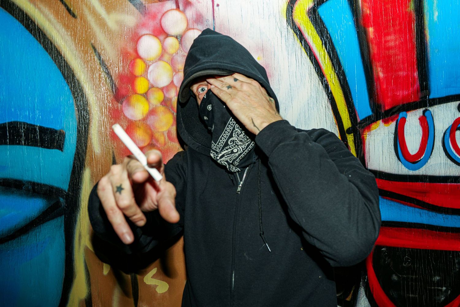 Person in black hoodie and bandana mask holding cigarette near colorful graffiti wall art.