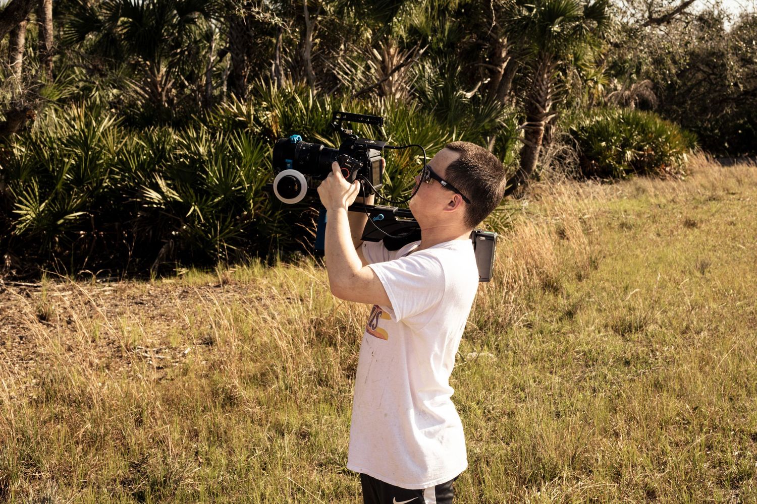 Filmmaker filming outdoors in a grassy field with dense trees using a professional video camera.