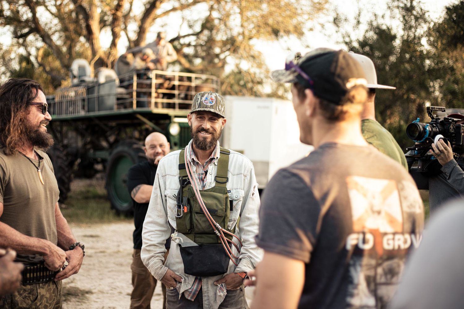 Men conversing on an outdoor film set with military vehicle and camera crew in background during golden hour.