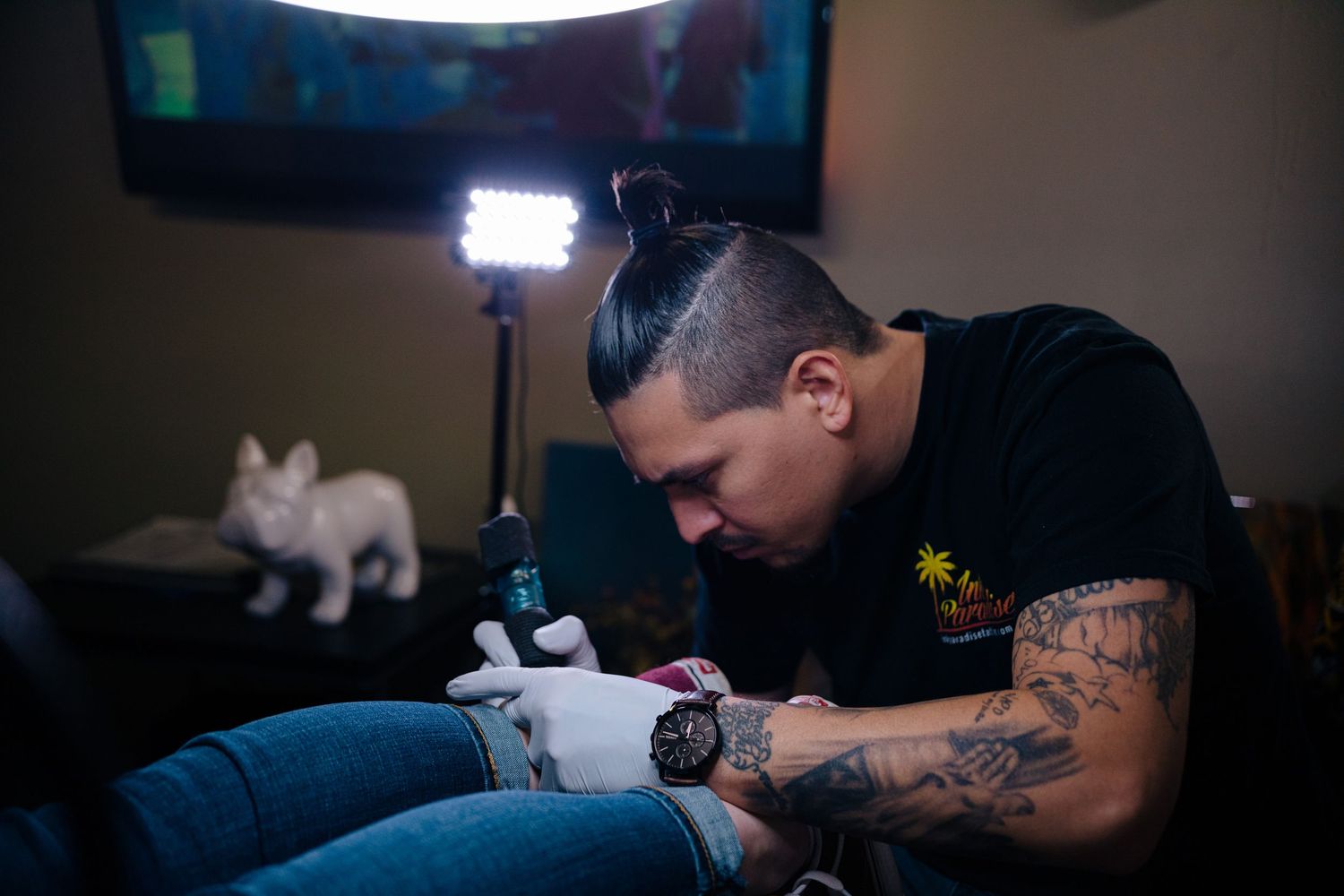 Tattoo artist with man bun and tattooed arms carefully inking a client, with a French Bulldog figurine in the background.