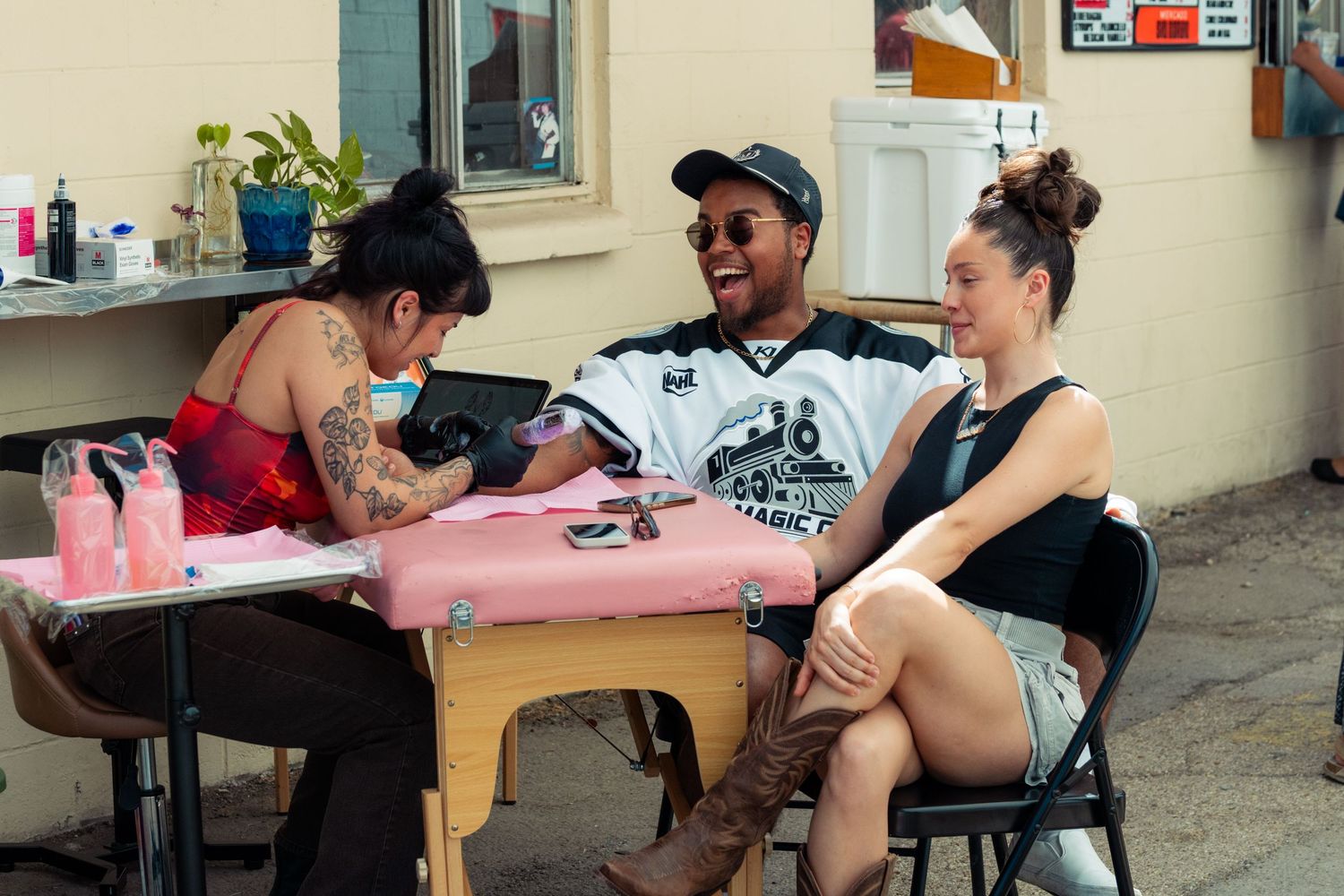 Tattoo artist inking a man's arm outdoors while a woman watches and they share laughs at a pink table.