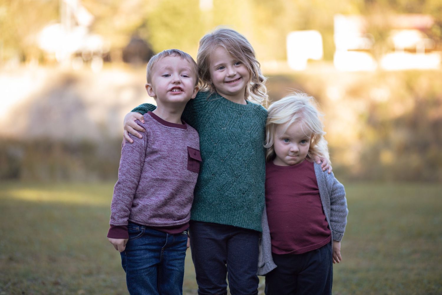 Three young siblings smiling and embracing each other outdoors in a grassy park.