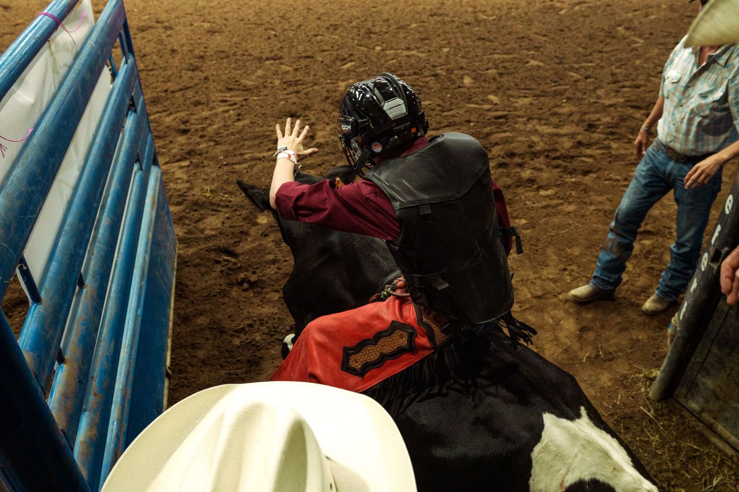 Bull rider in black helmet grips rope at rodeo chute, red vest visible, dirt arena background.