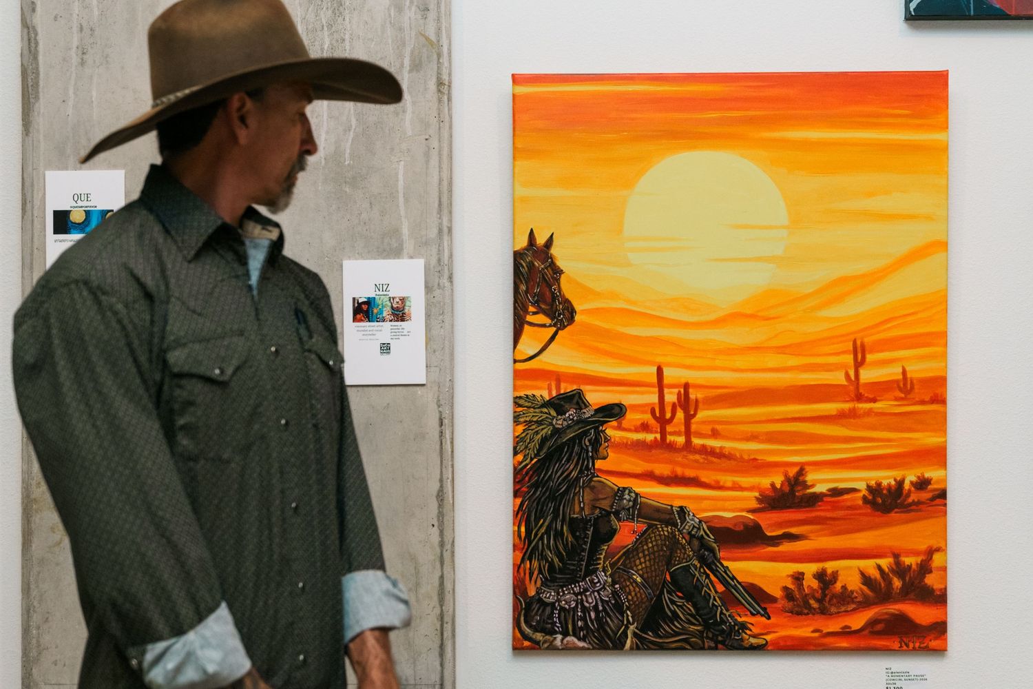 Man in cowboy hat stands beside vibrant orange desert sunset painting with horses at an art gallery exhibition.