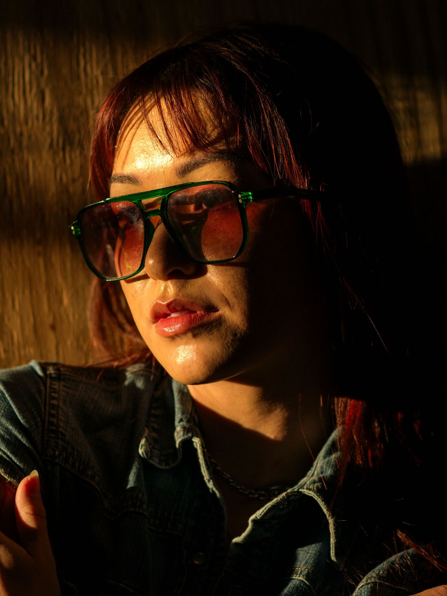 Young Asian woman with red hair wearing green oversized sunglasses in dramatic warm sunlight.