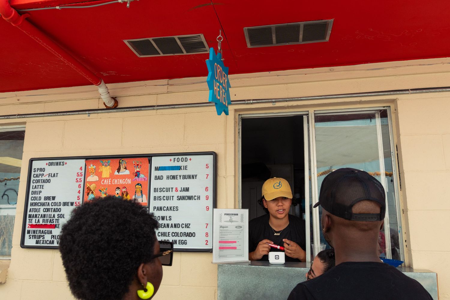 Worker serves customers at a red food stand window with a menu board visible outside.