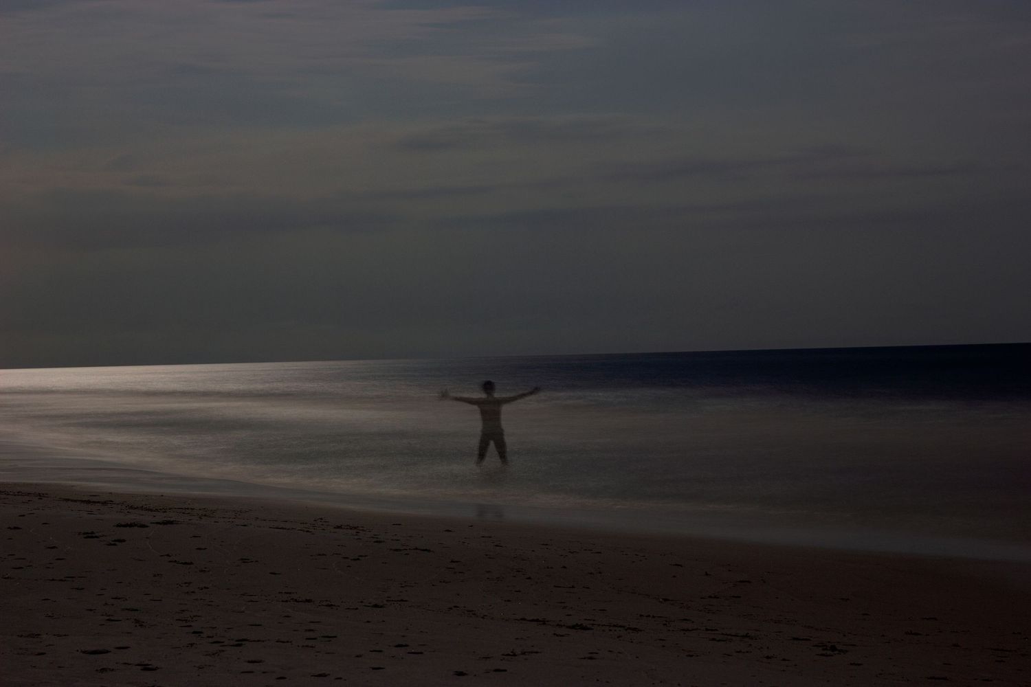 Silhouette of person standing on dark beach at dusk with arms outstretched, ocean waves reflecting moonlight.
