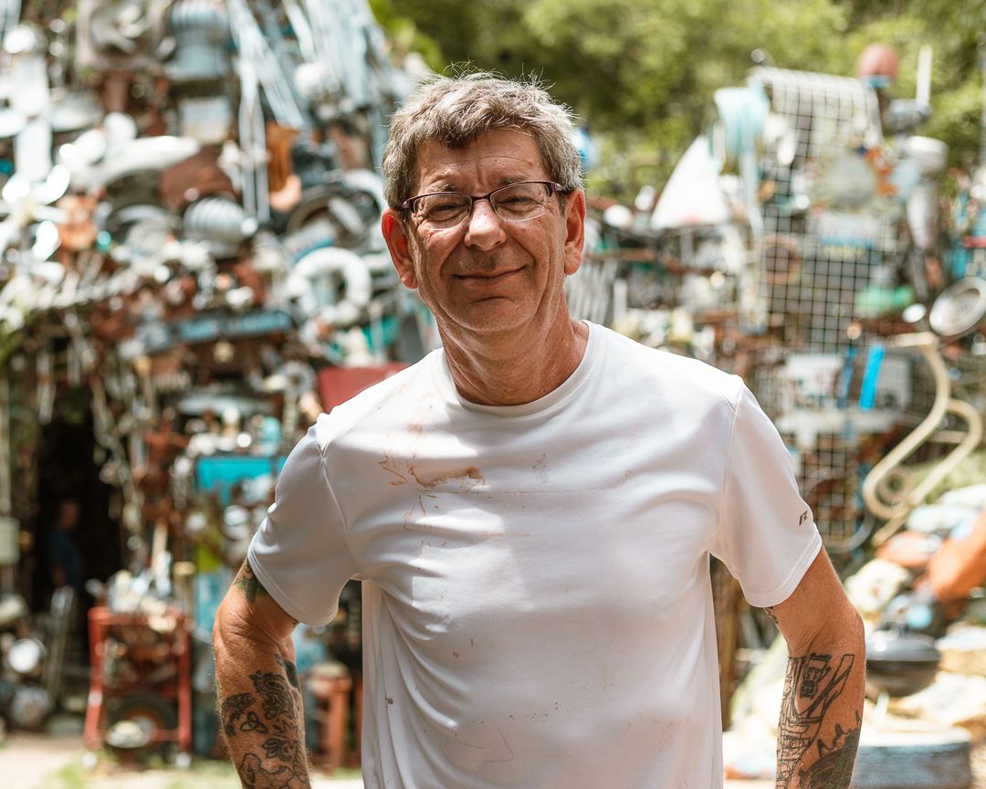 Tattooed man in white t-shirt smiling outdoors surrounded by colorful metal sculptures in a junkyard art installation.