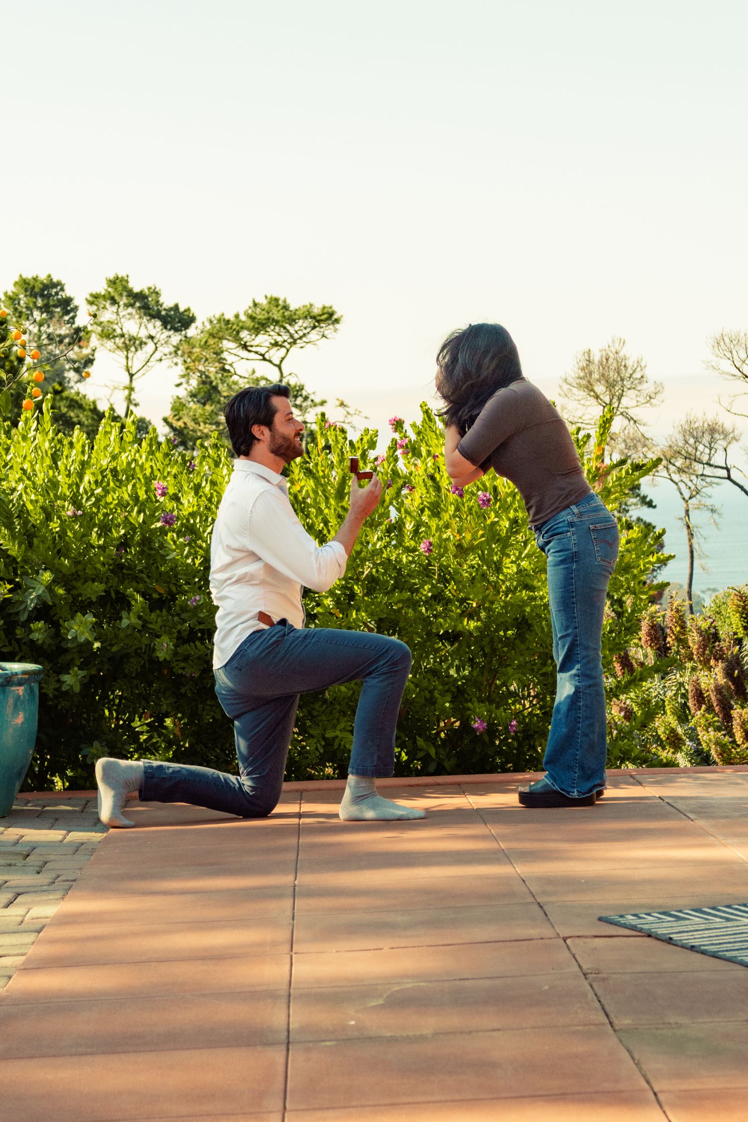 A man kneels on one knee proposing to a woman outdoors on a patio surrounded by lush green trees.