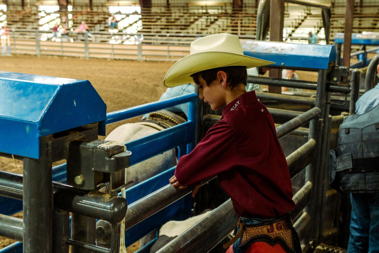Young cowboy in red shirt and hat leans on blue rodeo chute gate observing a bull at indoor arena.