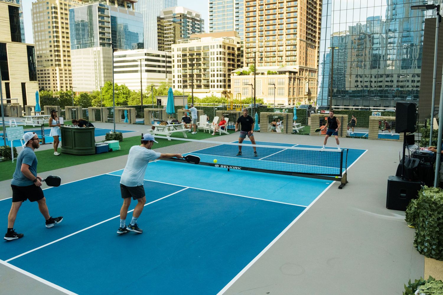 Players enjoy a pickleball match on a rooftop court with city skyscrapers in the background.