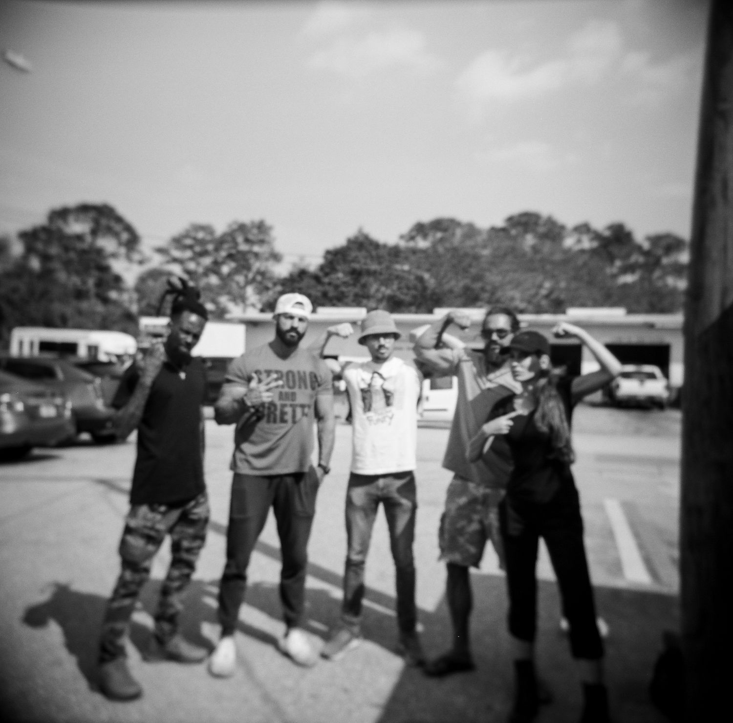 Five friends pose together in a parking lot, making gestures in a black and white outdoor photo.