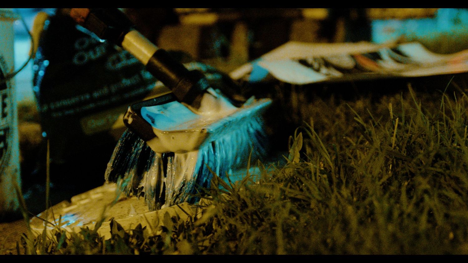 Close-up of a blue brush applying paint on grass outdoors at night.