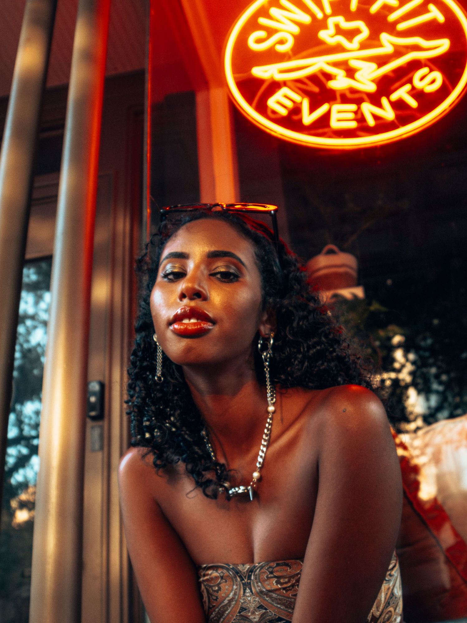 Elegant woman with curly hair posing under a glowing red neon events sign at night.