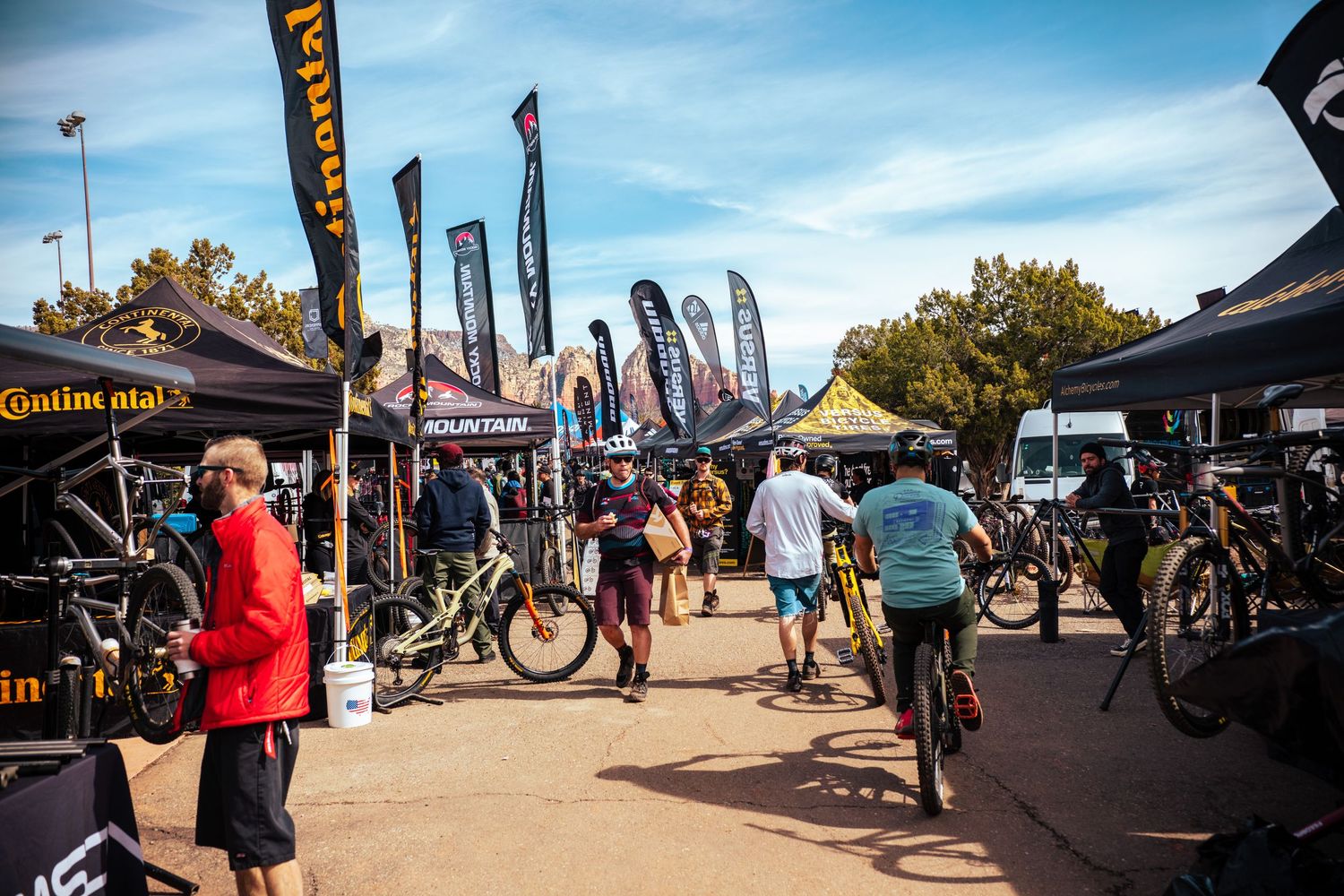 Busy outdoor mountain bike expo with vendor tents, banners, and cyclists browsing displays.