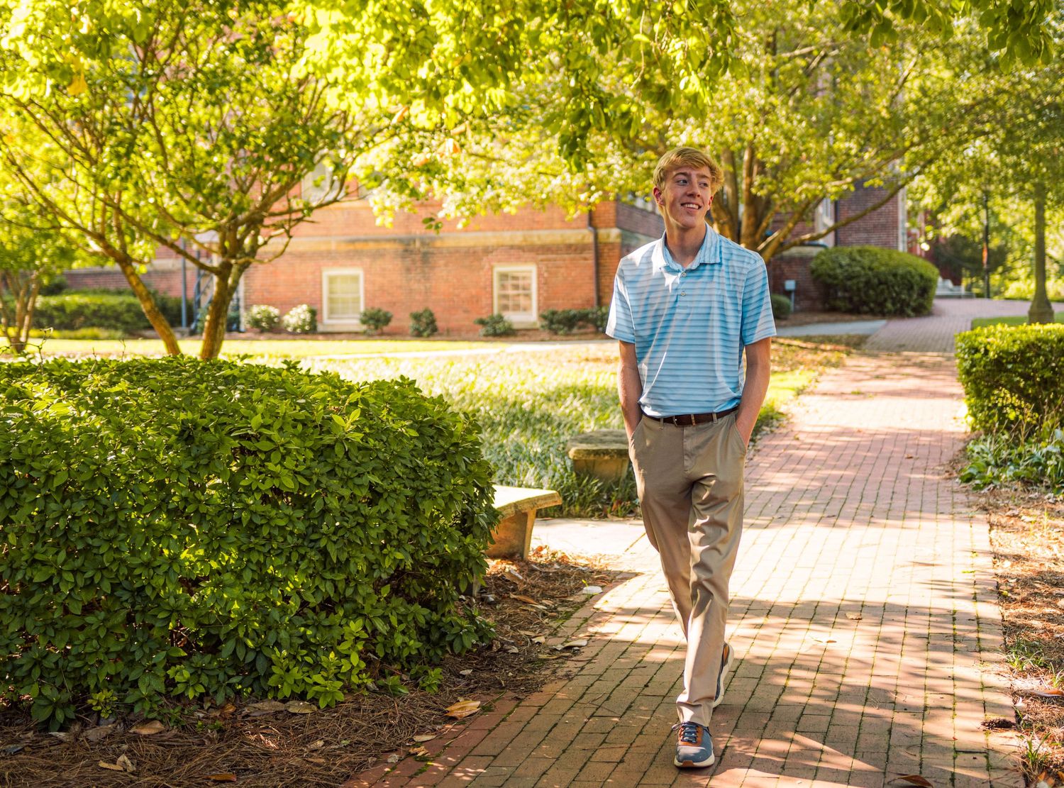 Young man in a light blue polo walks along a sunny brick pathway on a college campus.